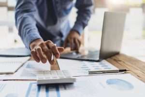 Un homme utilise une calculatrice sur un bureau avec des papiers et un ordinateur portable, calcule la taxe sur le profit immobilier